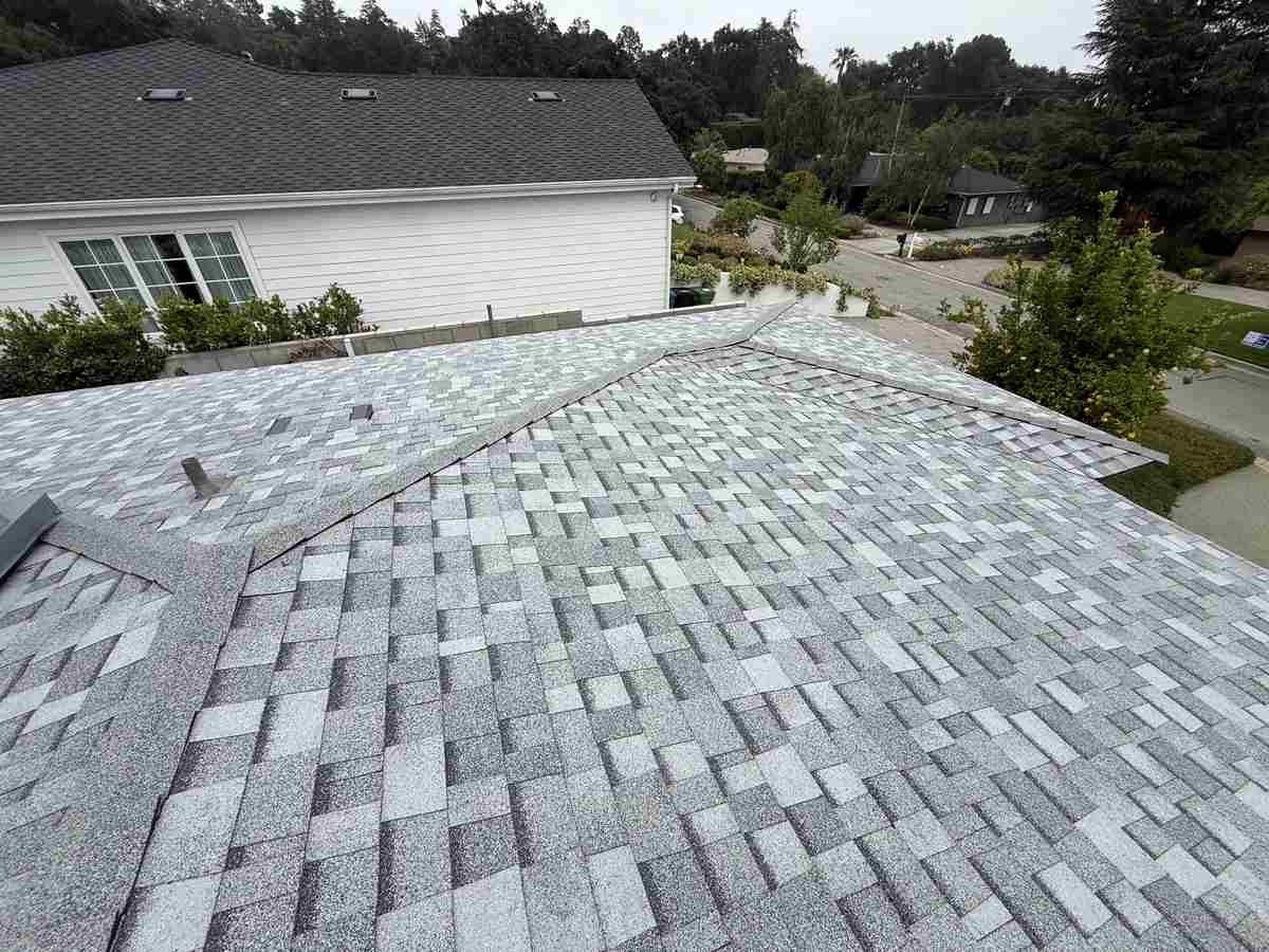 Light mottled gray shingle slopes and ridges seen from above, with vents, neighboring buildings and a street beyond